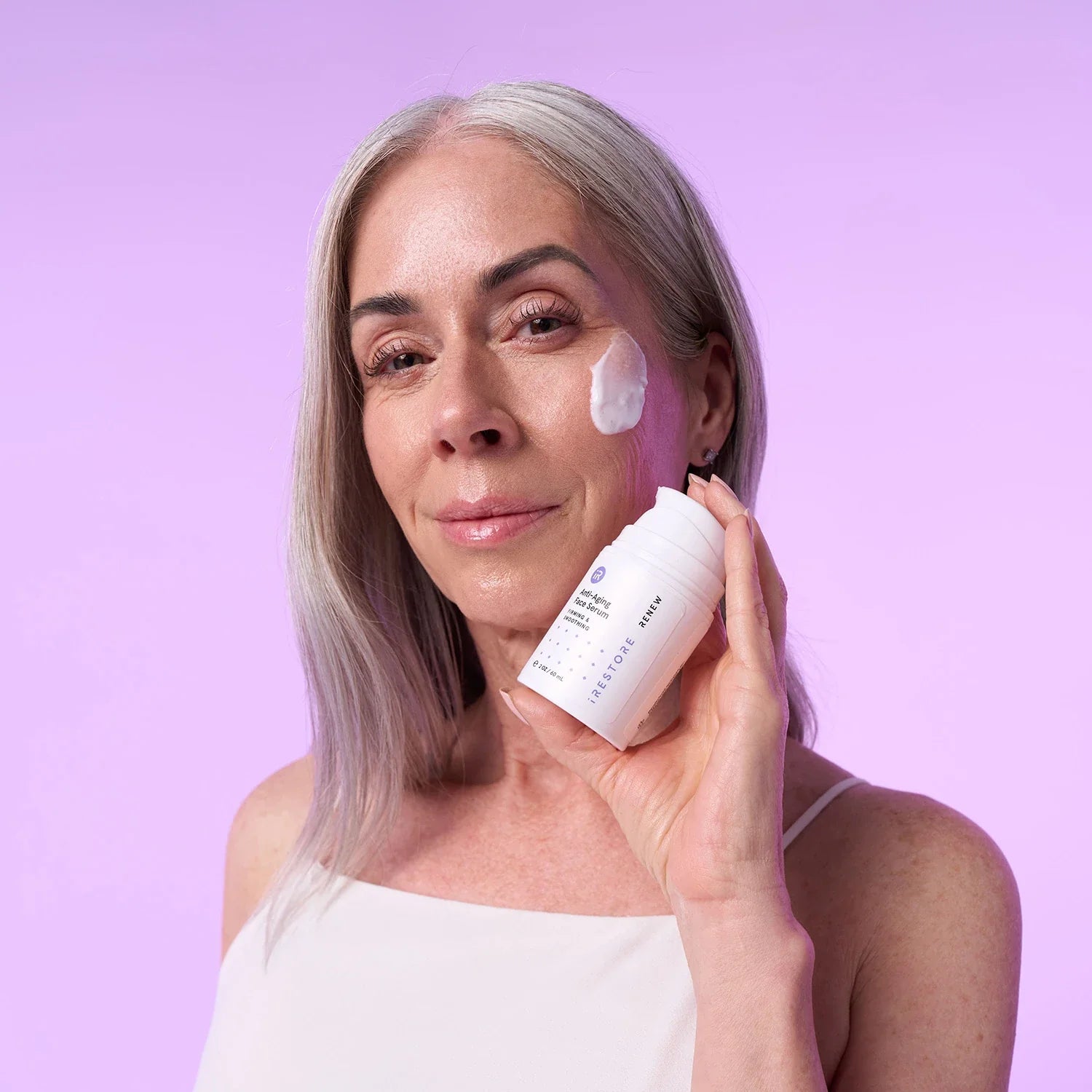 Mature woman with anti-aging face serum on her face while holding the bottle on her hand