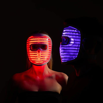 Front view of a woman wearing the iRESTORE LED face mask glowing red, standing beside a man in side view wearing the iRESTORE LED face mask glowing blue.