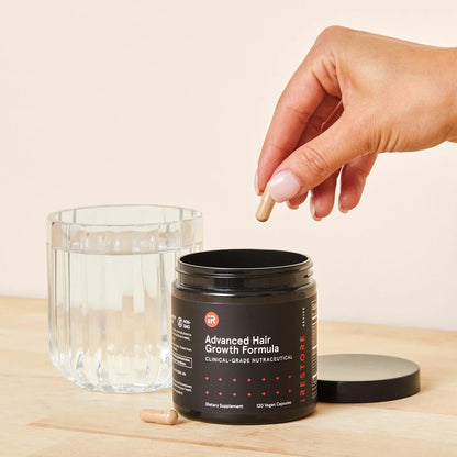 Image of a hand taking out a tablet in an Advanced Hair Growth Formula jar placed on a table with a glass of water beside it.