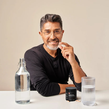 Image of a sitting man with Advanced Hair Growth Formula tablet in his hand. In front of him is a table with the cylindrical jar of the supplement placed on top of it with a glass and bottle of water beside it.